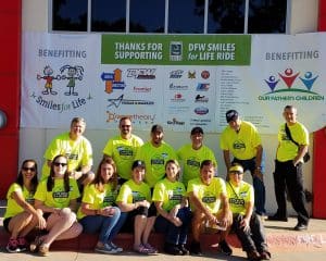 Smiling team in yellow shirts poses at charity event with sponsor banners.