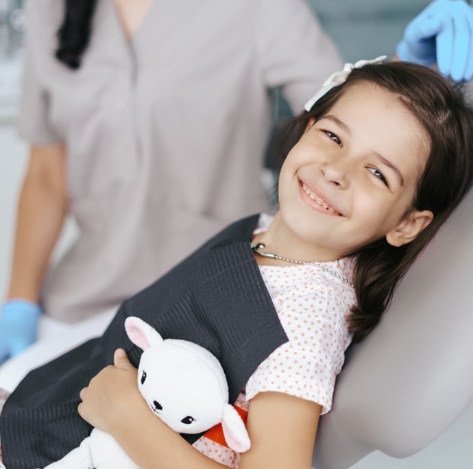 Young girl smiling in dental chair holding white stuffed animal toy at pediatric dental office.