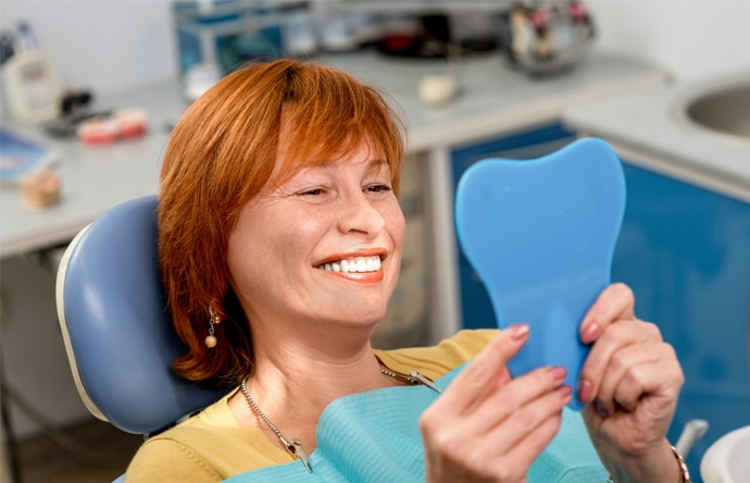 Woman with red hair smiling in dental office, holding blue mirror model.