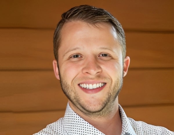 Professional headshot of smiling man in checkered shirt against wooden background for DFW Smiles dental practice.