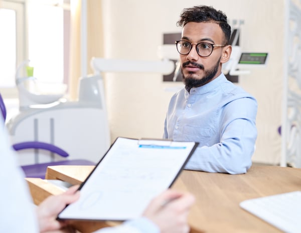 Male dentist in blue shirt holding clipboard in modern dental office treatment room.