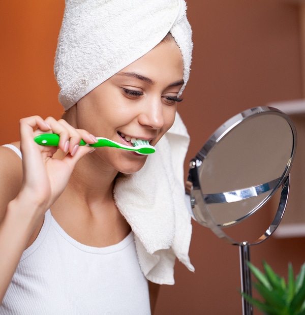 Woman brushing teeth at mirror with towel wrapped around head in bathroom setting.