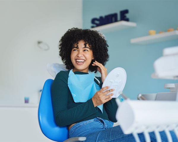 Woman with curly hair smiling in dental office chair holding white tooth model.