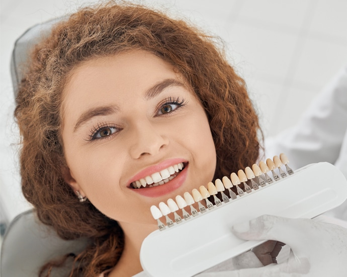 Woman smiling while holding a shade guide for dental whitening or color matching at DFW Smiles.