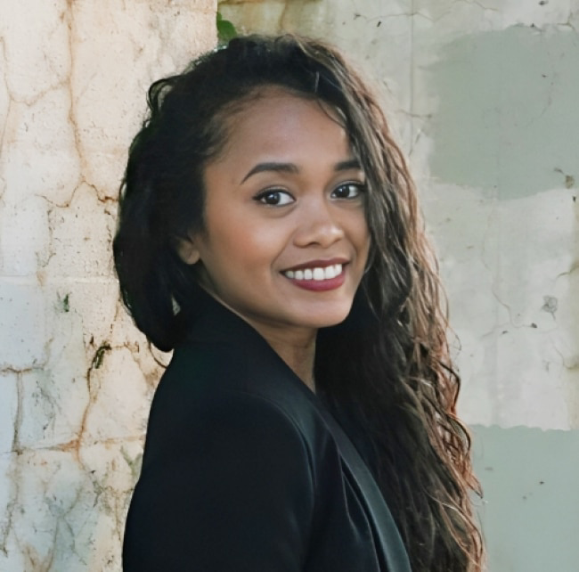 Profile portrait of a smiling young woman with long, dark, wavy hair. She is looking toward the camera, turned in profile against a weathered, light-colored stone wall. She is wearing a black blazer. The image has a vertical orientation with a thin, terracotta-colored border framing the top and bottom.